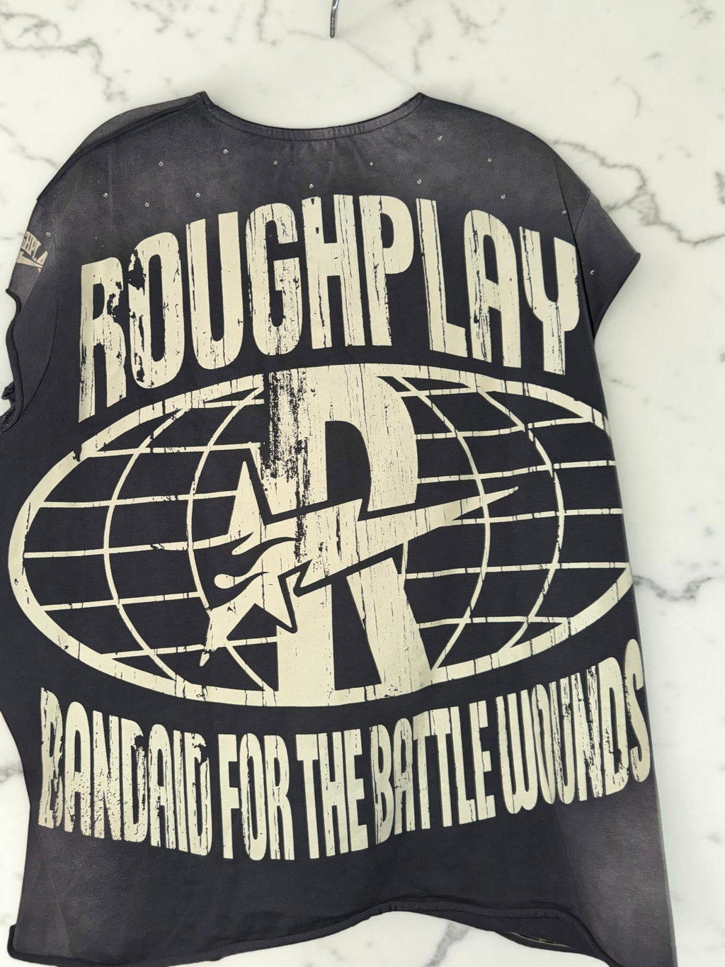 Back view of DRIPPING IN VINTY black graphic t-shirt with distressed ROUGH PLAY logo and white text on marble surface